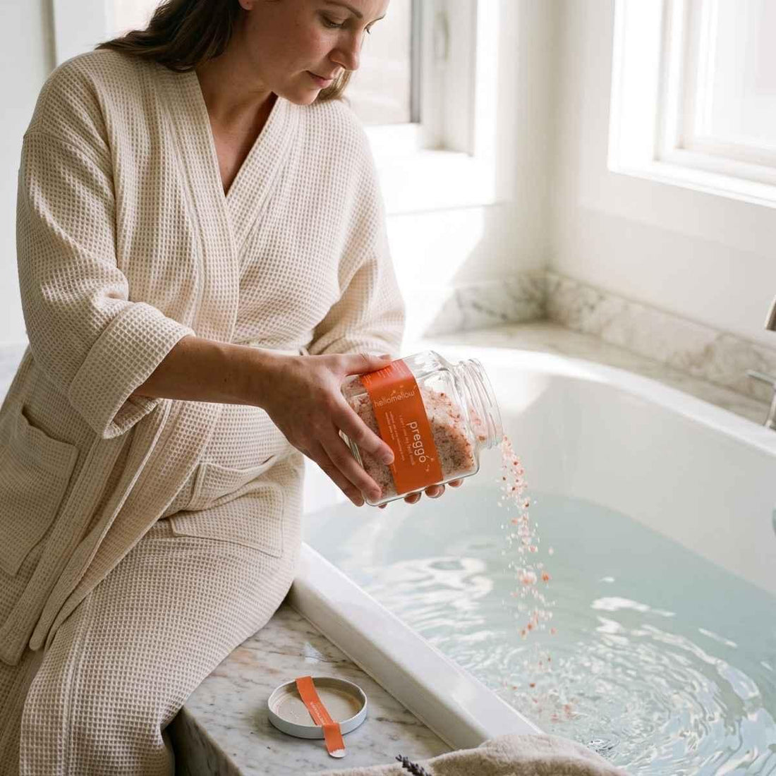 pregnant woman pouring i can’t see my feet soak into a bath beside a bright window.